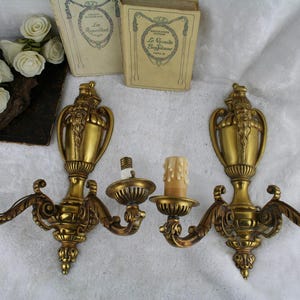 May include: A pair of matching gold-toned metal wall sconces, each featuring two curved arms with candle-style light sockets. The central vertical structure of each sconce is adorned with a detailed relief of a bearded face and an urn shape.