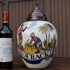 Pode incluir: Um grande vaso de cerâmica branca com tampa de bronze, apresentando uma cena pintada à mão de figuras e palmeiras. O vaso tem a palavra "TOEBACK" em uma faixa. Uma garrafa de vinho tinto está ao lado do vaso.