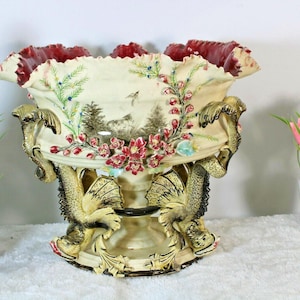 May include: An ornate, cream-colored ceramic vase with a ruffled, burgundy rim. The vase features a landscape scene with floral and dragon accents. A bottle of wine and a small potted plant are in the background.