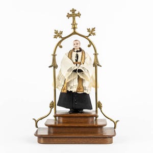 May include: A vintage religious figurine of a priest, dressed in a white and gold robe, standing on a wooden platform. The figurine is framed by a brass arch with a cross at the top. The priest's robe has a black skirt.