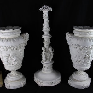 Op de afbeelding: Een set van drie witte decoratieve items: twee sierlijke urnen en een lange, gedetailleerde kandelaar. De urnen hebben reliëfsnijwerk van figuren en bloemmotieven. De kandelaar heeft een ronde basis en is versierd met cherubijnen.