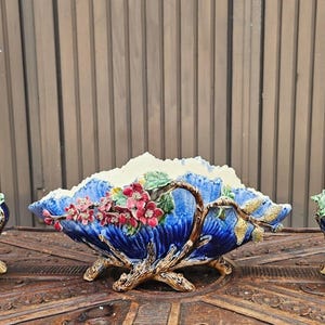 以下が含まれることがあります： ピンクの花、緑の葉、茶色の枝のレリーフが施された、2つの青い陶器製の背の高い花瓶と中央のボウルのセット。各ピースは溝のある質感で、枝のような形の脚の上に立っています。