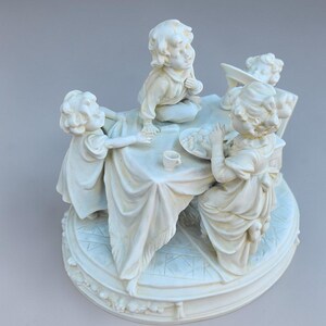 May include: A cream-colored figurine depicting four children gathered around a table. The children are dressed in vintage clothing and appear to be interacting with each other. The base of the figurine is round and decorated with floral patterns.