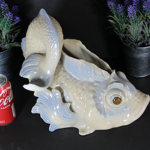 Peut inclure: Un pot de fleurs en céramique en forme de poisson, crème et bleu clair. Le poisson a des écailles, des nageoires et un grand œil détaillés. Le pot est à côté d'une canette de Coca-Cola rouge et de deux petits seaux en métal avec des plantes violettes.