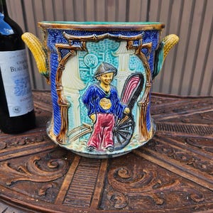 May include: A ceramic wine cooler featuring a relief design of a person pulling a rickshaw, accented with blue, green, and yellow tones. The vessel has two handles shaped like corn cobs and a textured surface with architectural motifs.