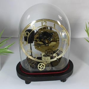 May include: An antique clock under a glass dome, revealing the internal mechanisms. The clock features a gold-coloured face and gears, set on a black base with a red trim. Two small potted plants with pink flowers are either side.