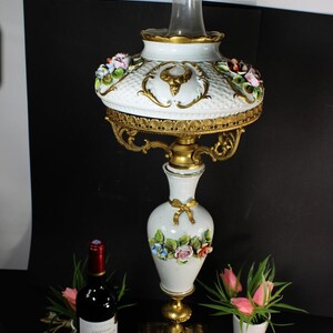 May include: A white and gold porcelain lamp with floral accents and a glass chimney. The lamp features a decorative base and ornate detailing. A bottle of red wine and a small potted plant are also visible.