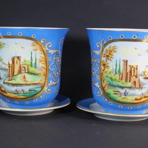 Może przedstawiać: Dwie niebieskie ceramiczne donice z pasującymi spodkami. Każda donica posiada ręcznie malowaną scenę krajobrazową w owalu ze złotymi obramowaniami. Donice mają rozszerzany kształt i są zdobione złotymi zawijasami.