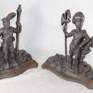 May include: A pair of dark brown cast metal bookends featuring figures of soldiers in traditional attire. One figure holds a tall spear, while the other holds a polearm with an axe head. Both figures stand on textured bases with rocky details.