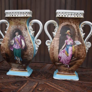 Puede incluir: Dos jarrones de cerámica con asas ornamentadas y bordes calados, con figuras pintadas a mano con vestimenta histórica. Un jarrón muestra a una persona con chaqueta morada y pantalones verdes, el otro a una persona con vestido rosa y abanico. Una botella de vino sirve de escala.