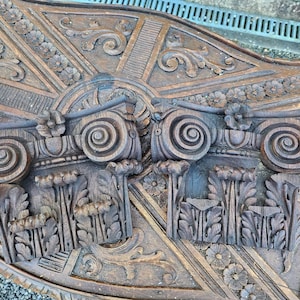 May include: A detailed, carved wooden panel featuring two classical columns with ornate scrollwork and floral accents. The panel has a warm, brown tone with visible wood grain and intricate relief carvings. The design includes stylized flowers and leaves.
