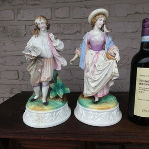 May include: Two porcelain figurines of a man and woman in period dress, alongside a bottle of red wine. The figurines stand on decorative bases. The woman wears a hat and holds a basket. The wine bottle has a white label.