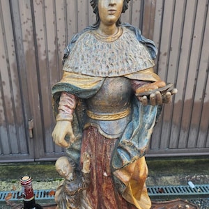 Puede incluir: Estatua de madera tallada de una figura vestida con una corona, sosteniendo un objeto pequeño. Una figura más pequeña está arrodillada en la base. Una botella de vino está al lado de la estatua.