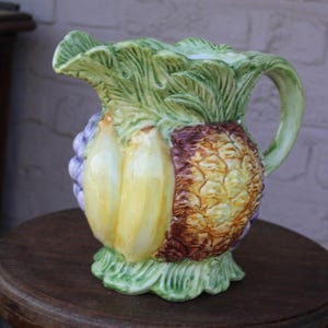 Peut inclure: Un pichet en céramique présentant un motif en relief tridimensionnel composé de divers fruits, dont un ananas brun texturé, des bananes jaunes et des raisins violets, encadrés par un feuillage vert aux bords avec une anse incurvée.