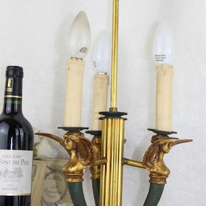 Könnte beinhalten: Ein gold-grüner Vintage-Kerzenleuchter mit drei Glühbirnen. Der Kerzenleuchter hat eine Mittelsäule und drei Arme, jeweils mit einer dekorativen Figur. Eine Flasche Rotwein ist links zu sehen.