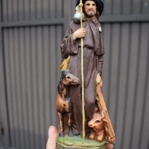 Puede incluir: Estatua marrón de un hombre con una túnica, sosteniendo un bastón y una bolsa. Un perro y un ternero están a sus pies. La estatua tiene un sombrero negro y una barba. La base de la estatua tiene la palabra "E GUIGON".