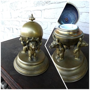 May include: A vintage brass container with a spherical lid and a base supported by three bear figures. The container is open, revealing a white interior. The brass has a warm, aged patina.