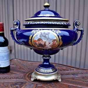 Könnte beinhalten: Eine tiefblaue Porzellanterrine mit Deckel, Goldverzierungen, kunstvollen Griffen und einem zentralen ovalen Medaillon, das eine klassische Szene mit Figuren und einem Pferd zeigt, neben einer Weinflasche als Größenvergleich.