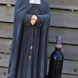 Puede incluir: Estatua de un hombre con túnicas oscuras, sosteniendo un rosario, con una botella de vino al lado. La estatua está sobre una pequeña mesa redonda azul oscuro. La estatua tiene un cuello blanco y una cabeza calva. La botella es negra con una tapa morada.