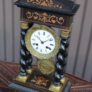 May include: A black mantel clock with gold-toned accents, featuring two twisted columns, a white circular face with black Roman numerals, and intricate gold-coloured metal detailing on the base, top, and surrounding the clock dial.