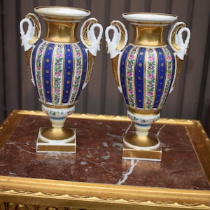 Puede incluir: Un par de jarrones de porcelana con un patrón de rayas azules y blancas, motivos florales, detalles dorados y asas en forma de cisne. Cada jarrón tiene un borde acampanado, un cuerpo cónico y una base cuadrada con acabados en oro.