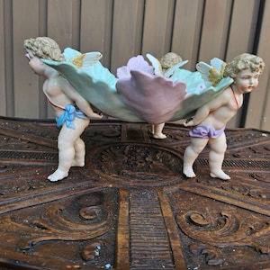 Puede incluir: Un centro de mesa decorativo de porcelana con tres figuras de querubines que sostienen un gran plato con forma de hoja festoneada. Los querubines tienen alas con detalles dorados y llevan paños de colores, con tonos pastel en verde y rosa.