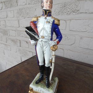 Puede incluir: Figura de cerámica de un oficial militar con uniforme azul, blanco y dorado, sosteniendo un sombrero y una espada. La figura se alza sobre una base rectangular con el nombre "LEPIC" inscrito. El uniforme del oficial presenta charreteras doradas y un cuello negro.