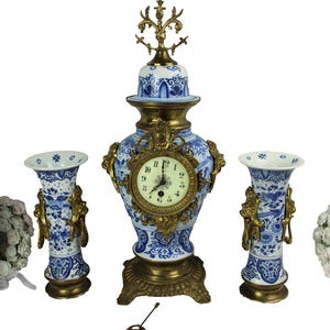 Könnte beinhalten: Eine Sammlung antiker Dekorationsartikel, darunter eine blau-weiße Porzellanuhr mit goldfarbenem Metallrahmen, zwei passende Vasen, eine Weinflasche und zwei Blumengirlanden. Das Zifferblatt der Uhr zeigt die Uhrzeit an.