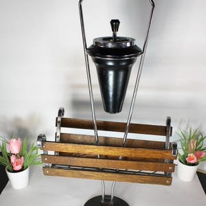 May include: A floor-standing smoking stand featuring a black conical ashtray with a chrome lid and handle, supported by a chrome frame. Below the ashtray is a rectangular wooden magazine rack with horizontal slats, all mounted on a circular base.