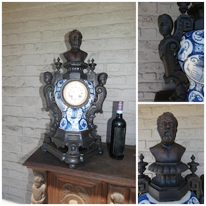 Könnte beinhalten: Eine antike schwarze Uhr mit einem weißen und blauen Porzellanzifferblatt und Akzenten. Die Uhr hat eine Büste eines Mannes an der Spitze und dekorative Figuren an den Seiten. Eine dunkle Weinflasche steht neben der Uhr auf einer Holzoberfläche.