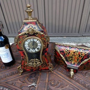 Puede incluir: Un reloj antiguo y una estantería de pared a juego con un diseño de carey rojo, negro y dorado. El reloj tiene una esfera blanca con números romanos negros. Una botella de vino tinto está a la izquierda.