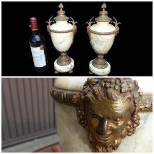 May include: A pair of white marble urns with gold-toned metal accents, featuring ornate handles and lids topped with finials. Each urn rests on a circular base. A wine bottle is placed next to the urns for scale. A close-up shows a gold face detail.