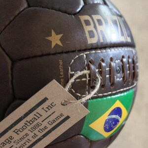 May include: A brown leather football with "BRAZIL" in gold lettering. The ball has a gold star and the Brazilian flag. A tag is attached with the text "Vintage Football Inc Since 1986 Spirit of the Game."