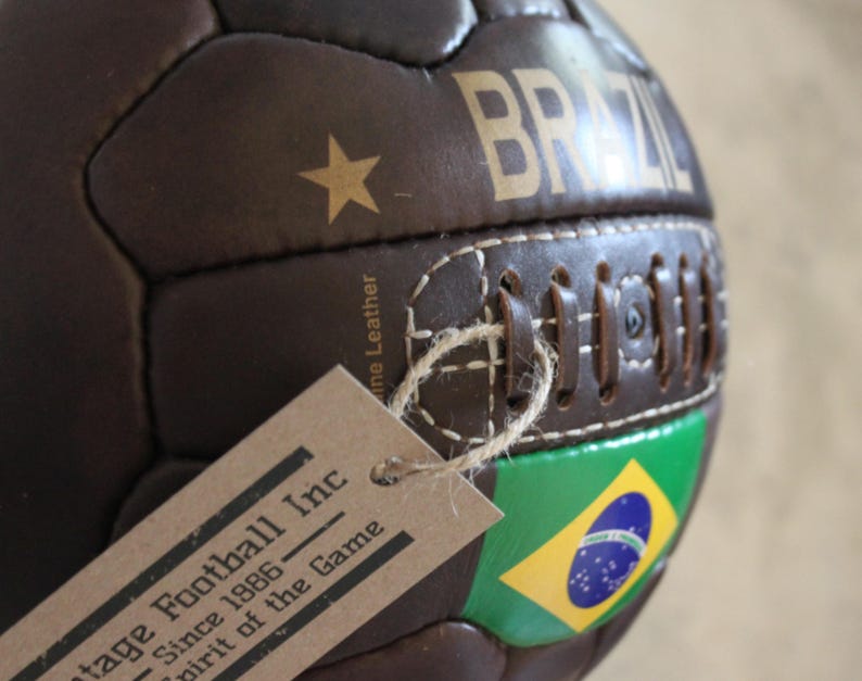 May include: A brown leather football with "BRAZIL" in gold lettering. The ball has a gold star and the Brazilian flag. A tag is attached with the text "Vintage Football Inc Since 1986 Spirit of the Game."