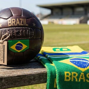 May include: A vintage brown leather football with "BRAZIL" printed on it, and a Brazilian flag patch. A matching green and yellow scarf with "BRASIL" is next to the ball. A tag reads "Vintage Football Inc. Since 1886".