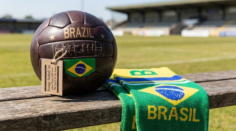 May include: A vintage brown leather football with "BRAZIL" printed on it, and a Brazilian flag patch. A matching green and yellow scarf with "BRASIL" is next to the ball. A tag reads "Vintage Football Inc. Since 1886".
