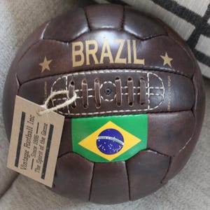 May include: A vintage brown leather football with "BRAZIL" in gold lettering and a Brazilian flag. The ball features a stitched design and a tag reading "Vintage Football Inc Since 1886 - The Spirit of the Game."
