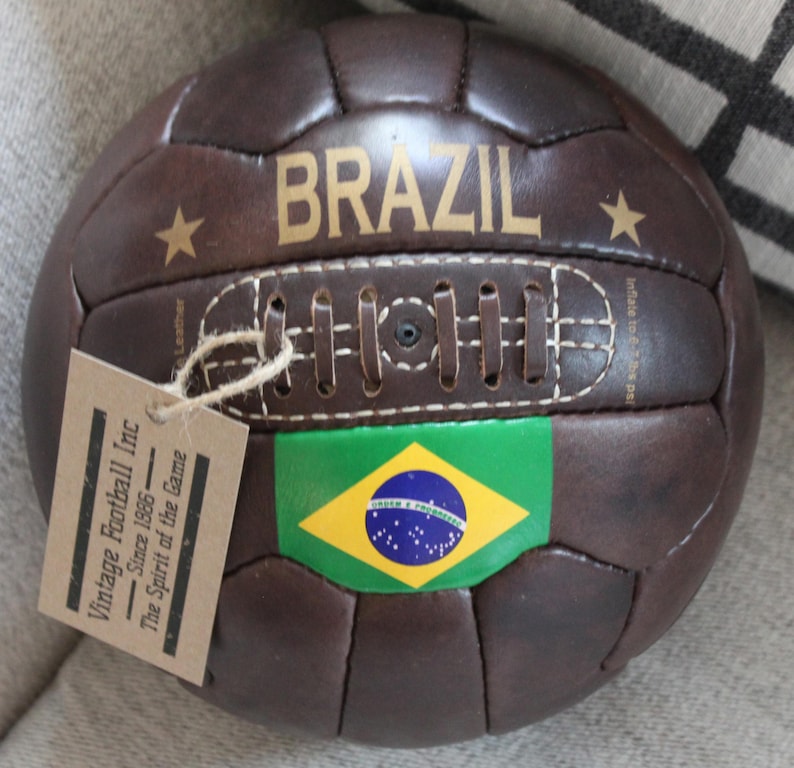 May include: A vintage brown leather football with "BRAZIL" in gold lettering and a Brazilian flag. The ball features a stitched design and a tag reading "Vintage Football Inc Since 1886 - The Spirit of the Game."