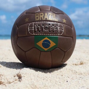 May include: A brown leather football with the word "BRAZIL" in gold lettering. The ball features the Brazilian flag and is resting on a sandy beach with a blue sky in the background.