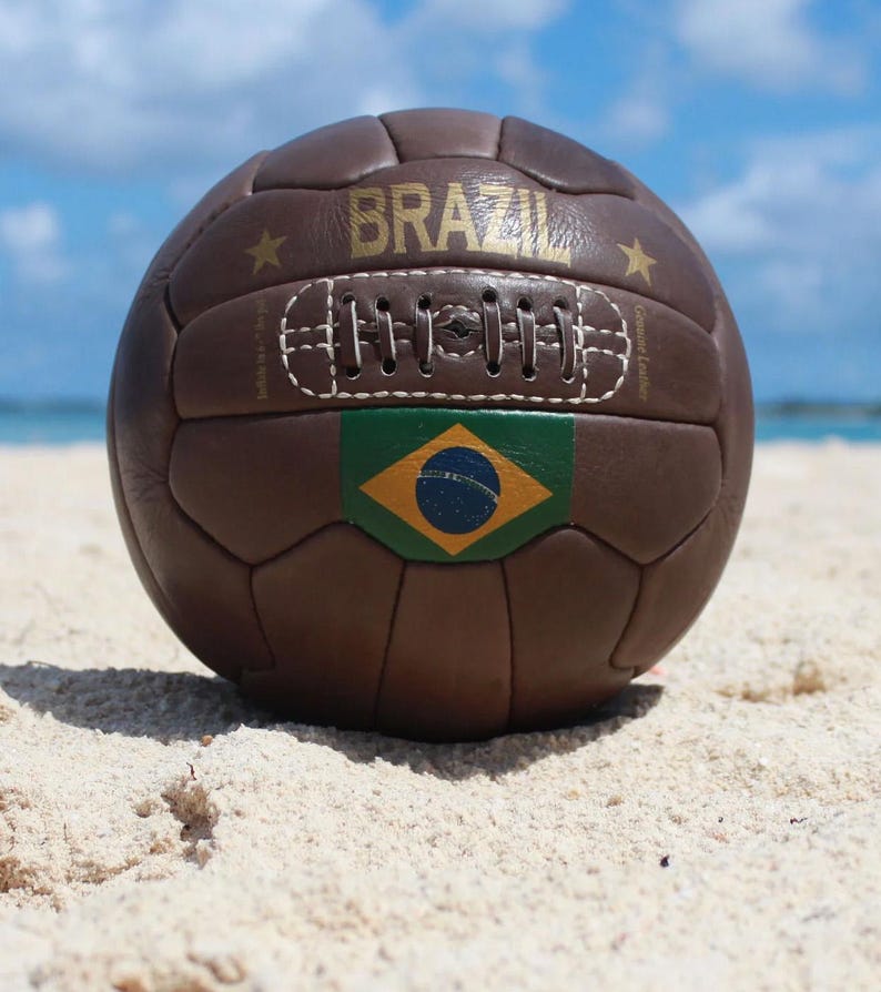 May include: A brown leather football with the word "BRAZIL" in gold lettering. The ball features the Brazilian flag and is resting on a sandy beach with a blue sky in the background.
