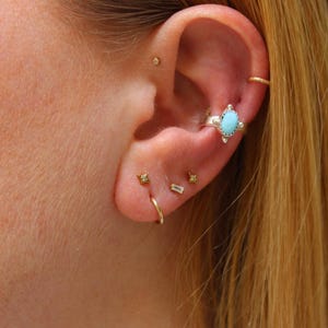May include: Close-up of an ear with various earrings. A small gold hoop, a turquoise and silver ear cuff, and several small stud earrings with clear stones are visible. The ear is set against a backdrop of blonde hair.