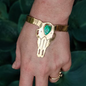 May include: A gold-colored cuff bracelet featuring a stylized skull design with a teardrop-shaped, green gemstone at the center. The bracelet has a textured surface and is worn on a wrist.