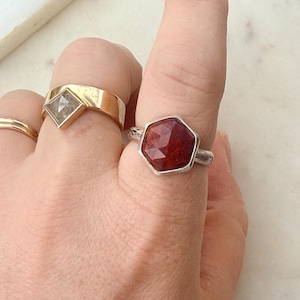May include: A hand wearing three rings. The first ring is gold with a clear, diamond-shaped stone. The second ring is a simple gold band. The third ring is silver with a faceted, hexagonal, red gemstone.