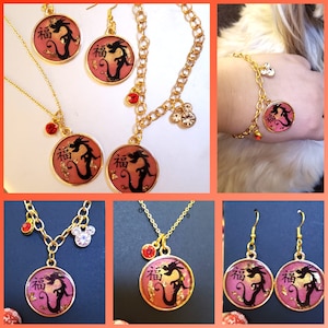 Puede incluir: Un conjunto de joyas de oro con un colgante de dragón rojo y dorado con el carácter chino para "buena suerte". El conjunto incluye un collar, pendientes, pulsera y un amuleto.