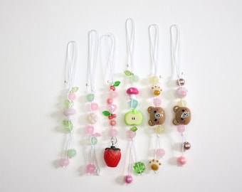 May include: A collection of six beaded charms with white cords. The charms feature various designs, including fruits, flowers, and bear faces. The beads are in shades of pink, green, and white, creating a playful and colorful aesthetic.