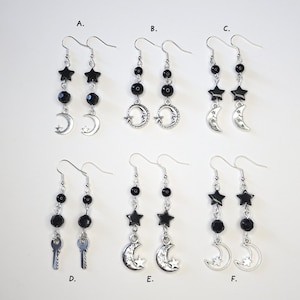 May include: Six pairs of dangle earrings are shown. Each pair has black beads, star, moon, or key charms. The earrings are silver-toned and hang from a hook. The earrings are displayed against a white background.