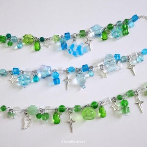 Könnte beinhalten: Drei mit A, B und C gekennzeichnete Bettelarmbänder mit verschiedenen Perlen in Blau- und Grüntönen. Die Armbänder enthalten silberfarbene Stern- und Kreuzanhänger sowie durchscheinende Glasperlen in verschiedenen Formen wie Blumen und Kugeln.