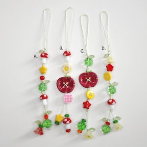 May include: Four whimsical beaded charms with various designs. Each charm features a loop for hanging and includes elements like mushrooms, stars, apples, and floral accents in red, yellow, green, and white. The charms are labeled A, B, C, and D.