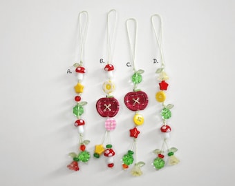 May include: Four whimsical beaded charms with various designs. Each charm features a loop for hanging and includes elements like mushrooms, stars, apples, and floral accents in red, yellow, green, and white. The charms are labeled A, B, C, and D.