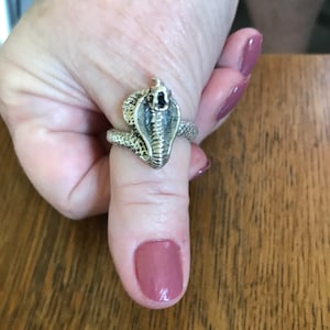 May include: A silver ring with a detailed cobra design. The ring is worn on a finger with pink nail polish.